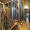 Two groups of organ pipes inside the Bigelow organ chamber at Saint Ambrose Church, with stained glass window seen through door.